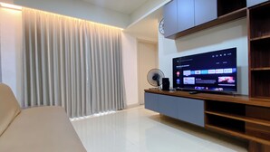 A modern living room with a flat-screen TV, a wooden entertainment unit, a fan, and a large window with vertical blinds. - Well Furnished And Comfortable 2Br Meikarta Apartment (Cikarang)