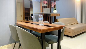 A modern living room with a wooden dining table, two chairs, a beige sofa, and a built-in shelving unit with decorative items. - Well Furnished And Comfortable 2Br Meikarta Apartment (Cikarang)