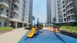 A playground with a yellow slide and climbing structures in a residential area with high-rise buildings. - Good And Simply Studio Tokyo Riverside Pik 2 Apartment (Teluknaga)