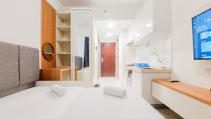 A modern hotel room with a bed, a grey sofa, a wooden shelf, a TV, and a bathroom with a sink and mirror. - Cozy Living Studio Sky House Bsd Apartment Near Aeon Mall (Pagedangan)