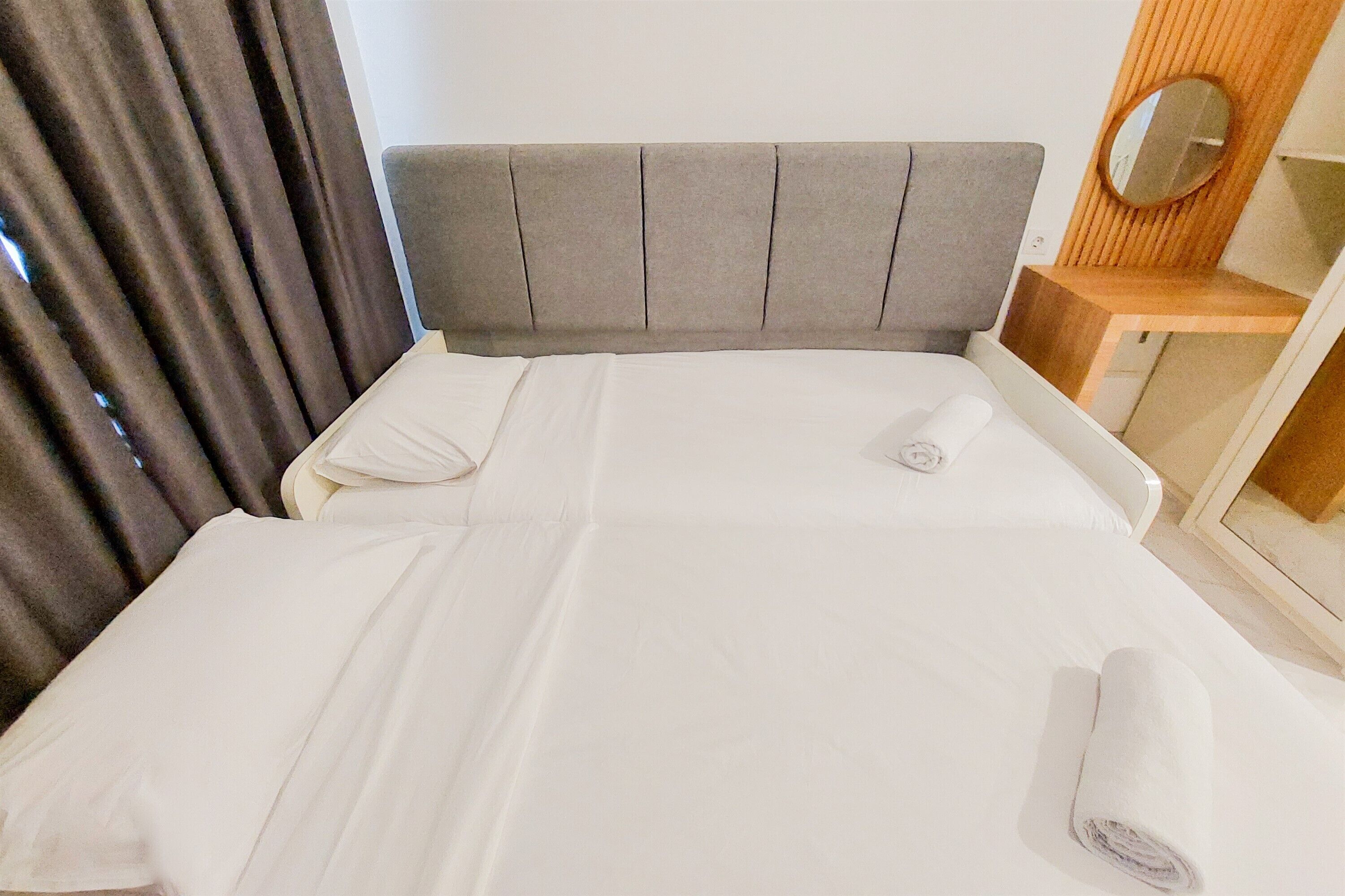 A neatly made bed with white linens, a grey upholstered headboard, and a wooden side table with a mirror.