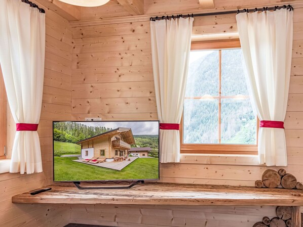 A wooden cabin interior with a TV displaying a house, curtains, and a window with a mountain view. - Glockner by Interhome (Heiligenblut)