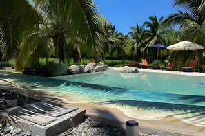 A clear swimming pool surrounded by rocks and palm trees, with lounge chairs and umbrellas in the background.
