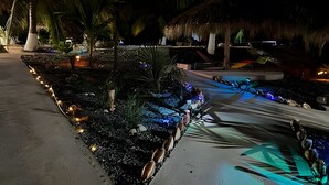 A nighttime resort area with a pathway lined with decorative lights, palm trees, and a thatched-roof structure.