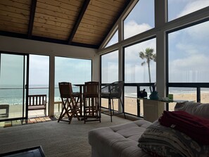 A spacious room with large windows overlooking a beach, featuring a wooden ceiling, a sofa, and a table with chairs.