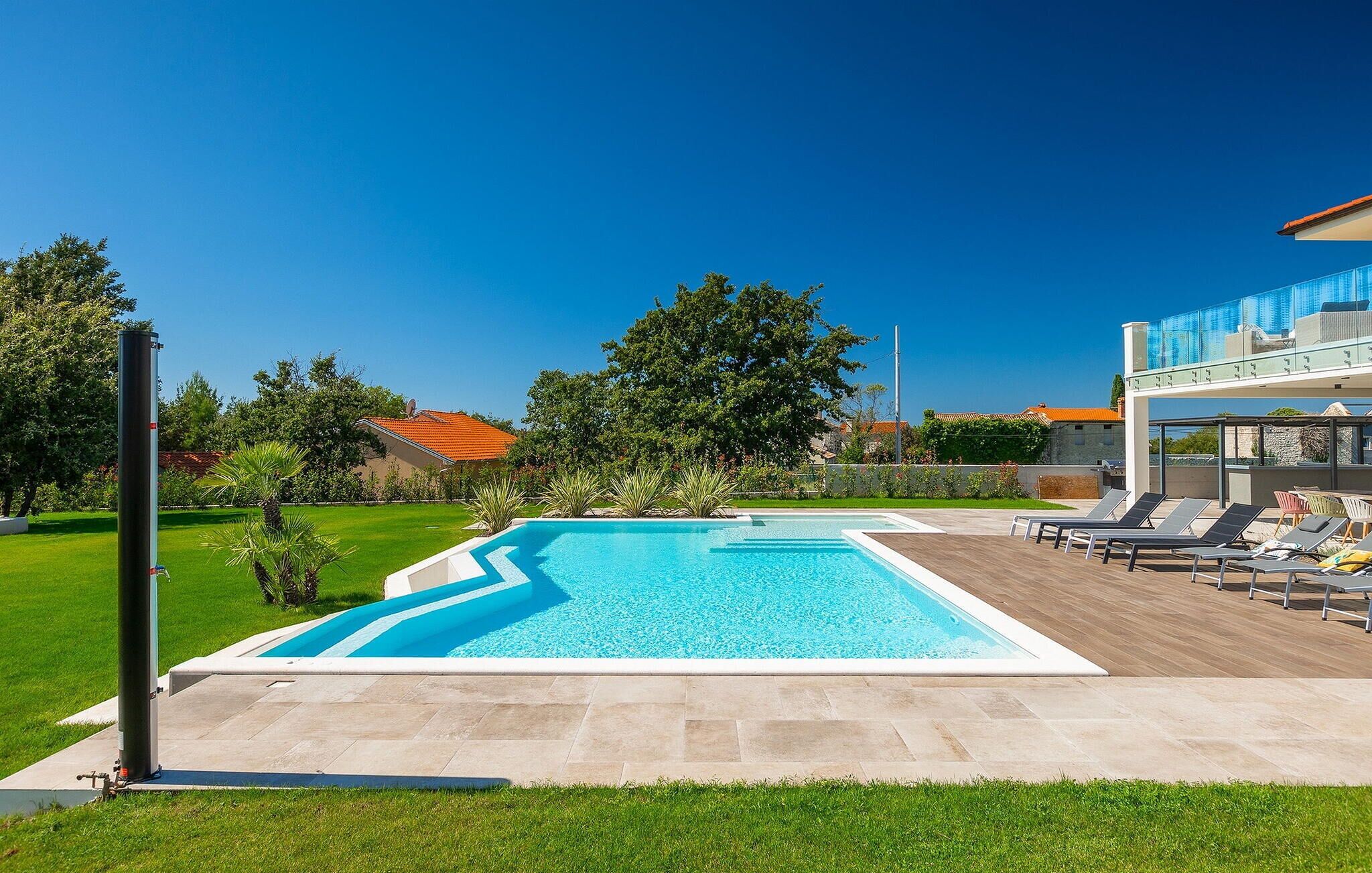 Ein Schwimmbecken mit Holzterrasse und Liegestühlen, umgeben von Grünpflanzen und blauem Himmel.