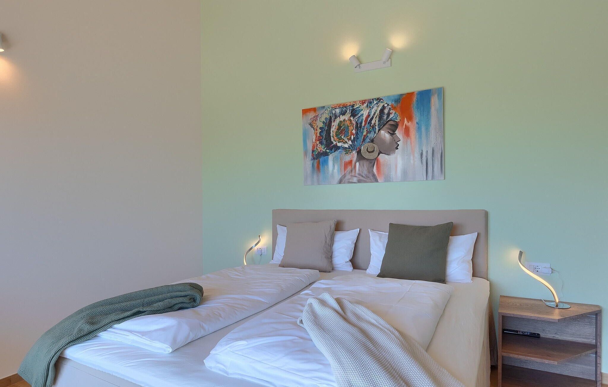 A neatly made bed with white linens and a green throw blanket, a headboard with multiple pillows, a wall-mounted light fixture, and a painting above the bed.