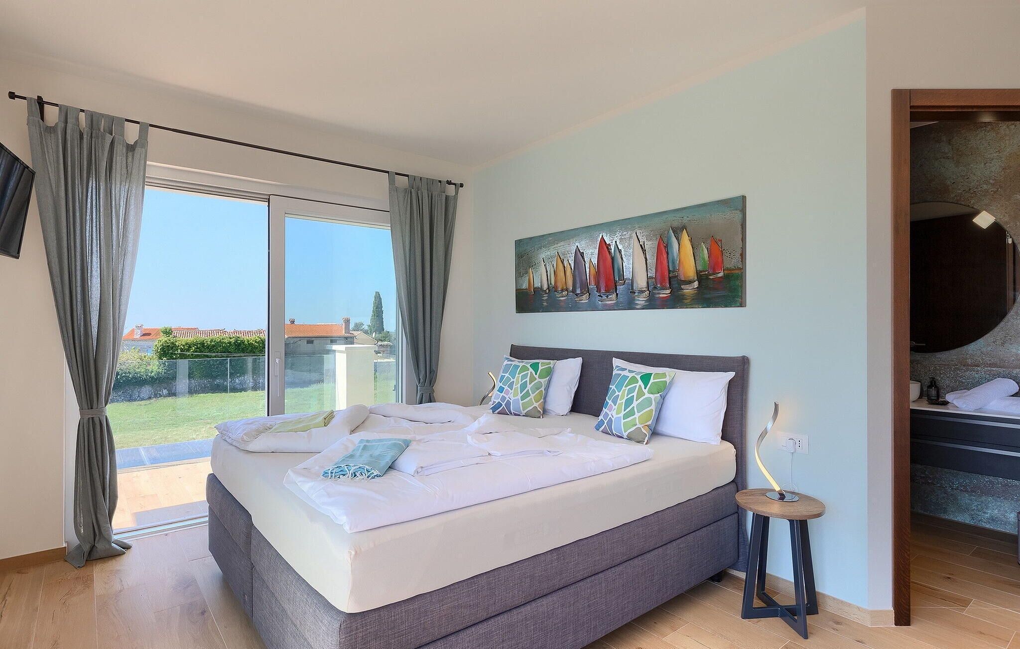 A bedroom with a large bed, a painting of sailboats, a small stool, and a view of the outdoors.