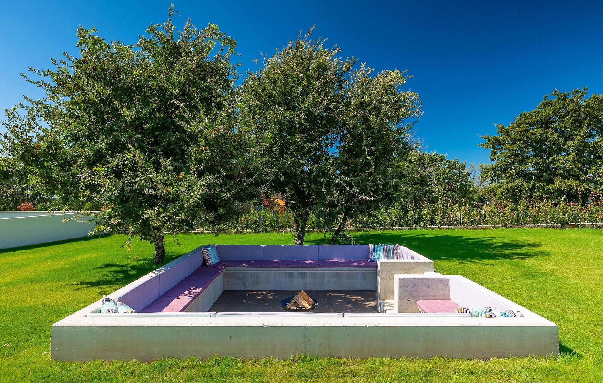Un espace extérieur aménagé en béton, avec des bancs et un foyer, entouré d’arbres et d’herbe.