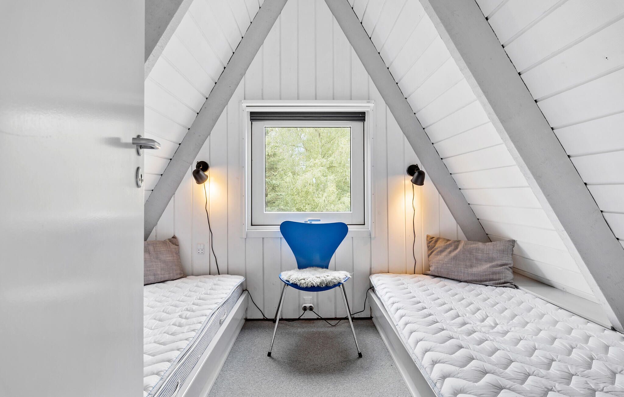 A small attic bedroom with a sloped ceiling, two single beds, a blue chair, and two wall-mounted lamps.