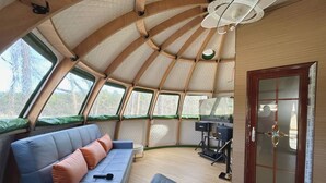 A spacious, high-ceilinged room with a wooden floor, a sofa with cushions, a small table, and a large window. - Countryside Getaway Green UniqueDomeA/serene farm retreat (Snow Camp)