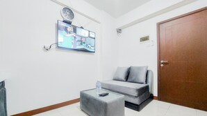 A living room with a grey sofa, a glass coffee table with a remote control and a water bottle, a mounted TV displaying a video call, and a wooden door. - Good Price And Homey 2Br At Loftvilles City Apartment (South Tangerang)