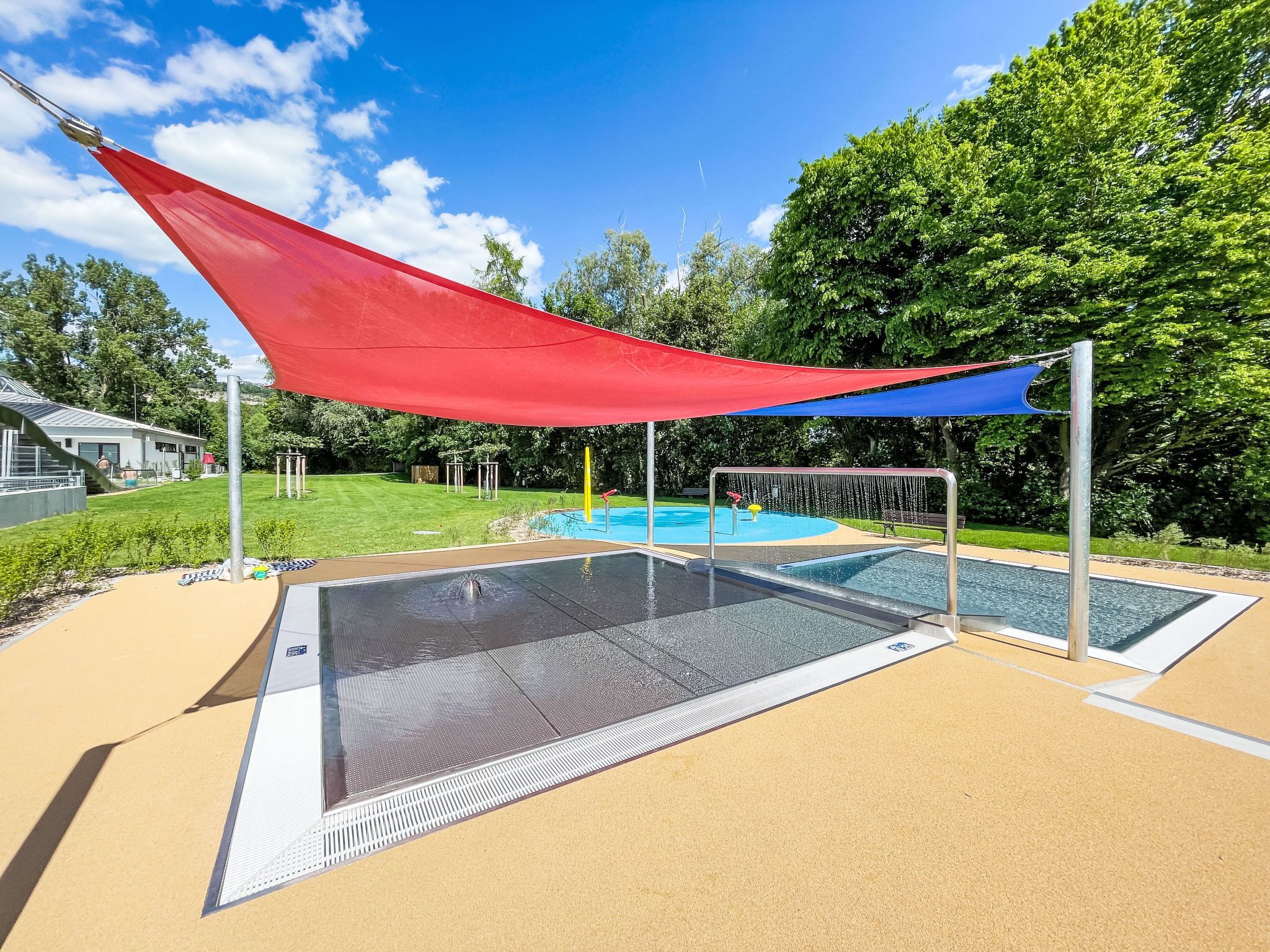 Ein Poolbereich mit rotem Sonnensegel, blauem Sonnensegel und einem Wasserspiel.