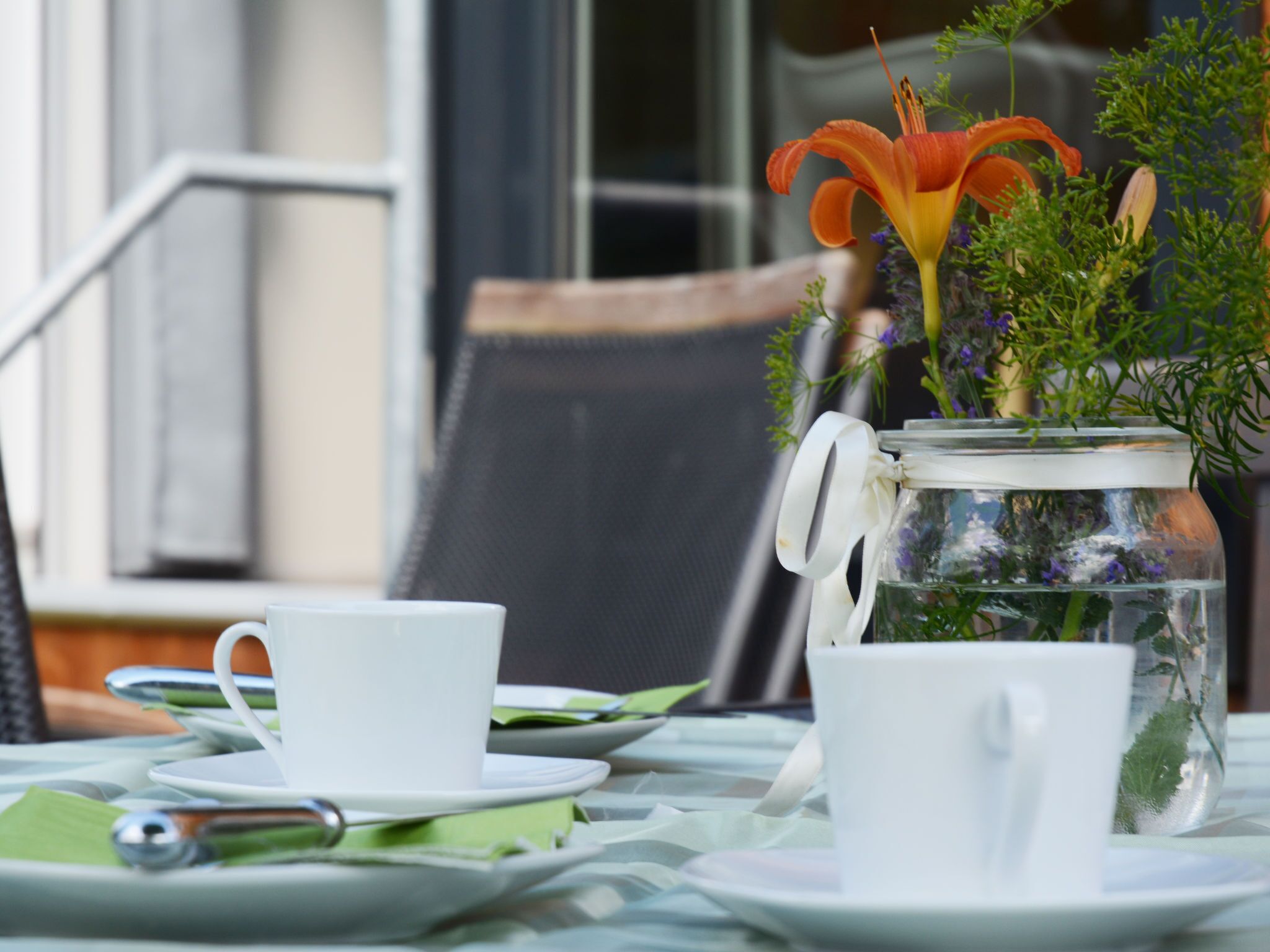 Una mesa servida con tazas y platillos blancos, una cuchara de metal y un frasco de vidrio con un lazo blanco.