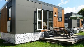 Een modern, compact hotel met een grijze gevel, houten details en een klein terras met een tafel en stoelen.