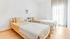 Un dormitorio con dos camas, una mesita de noche con lámpara y una ventana con cortinas translúcidas.