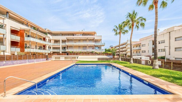 Una piscina rodeada por un edificio de varios pisos con balcones, palmeras y un cielo despejado.