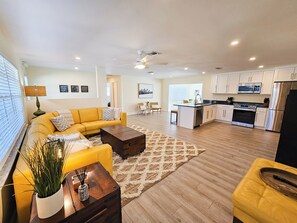 Ein modernes Wohnzimmer mit einem gelben Sofa, einem Holztisch und einer Küchenzeile mit weißen Schränken und Edelstahlgeräten.