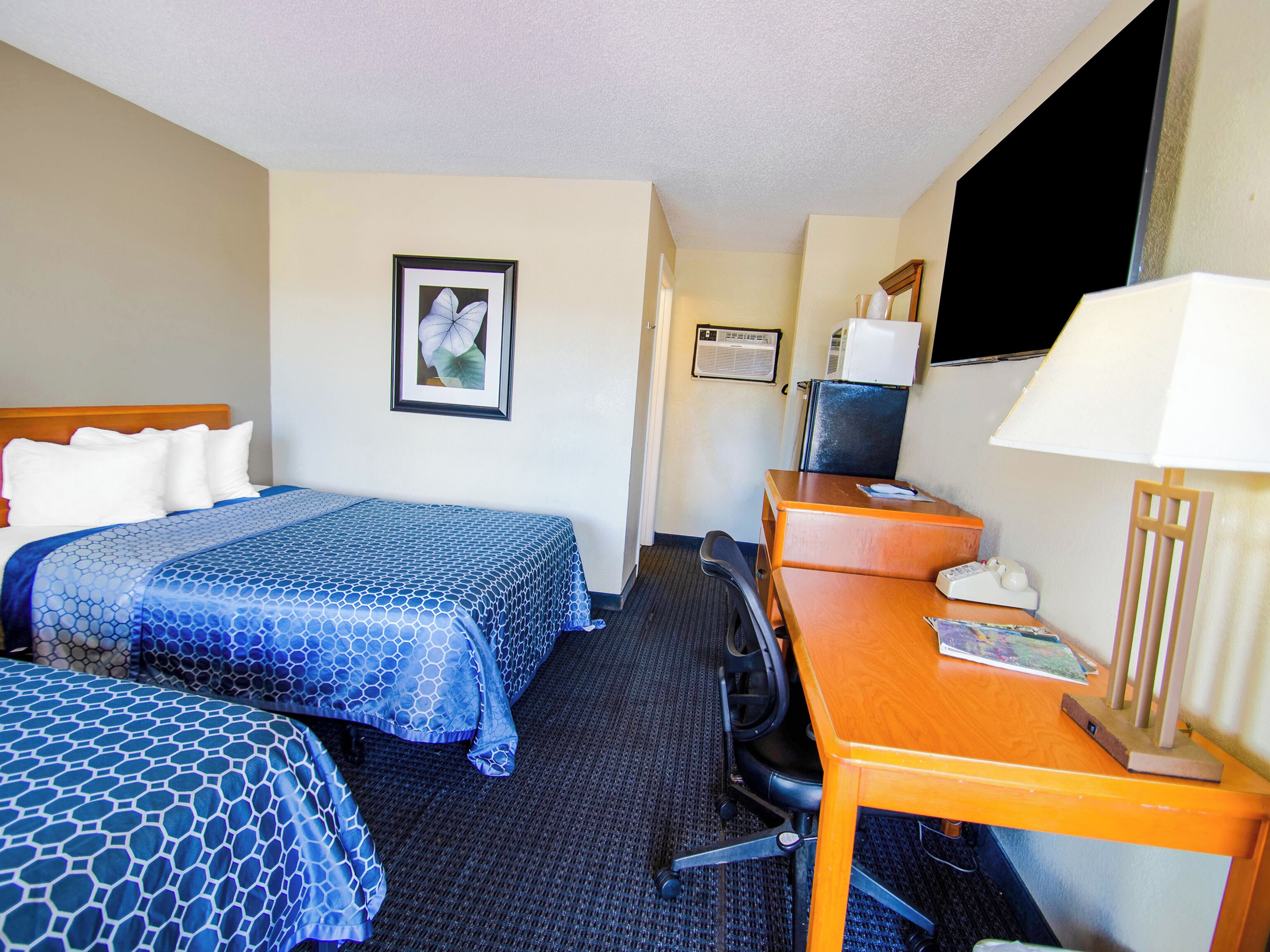 A hotel room with two beds, a desk, a chair, a lamp, a picture on the wall, and an air conditioning unit.