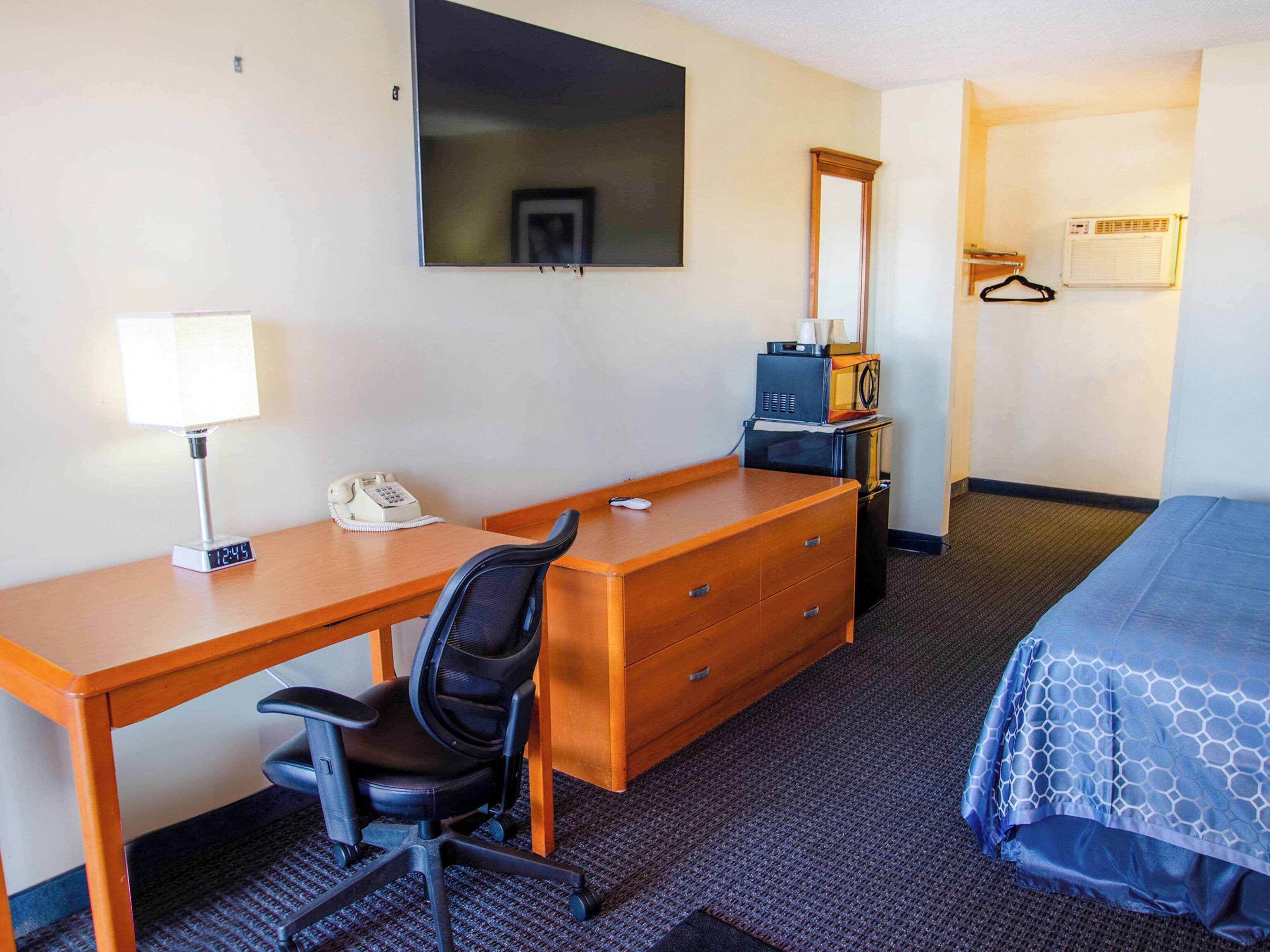 A hotel room with a wooden desk, a black office chair, a bed with a blue patterned bedspread, a television mounted on the wall, a microwave, and a small heater.