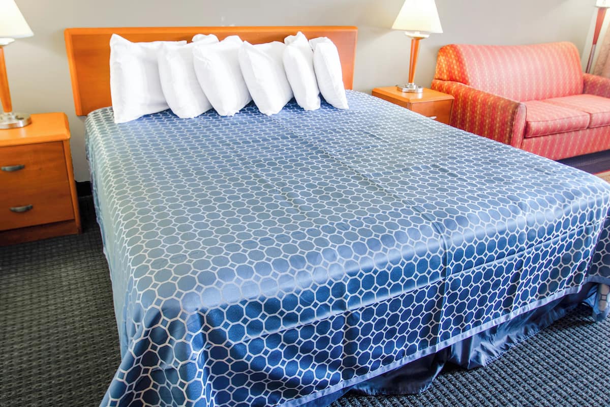 Habitación de hotel con una cama grande, cabecero de madera, dos mesitas de noche con lámparas, un sillón rojo y una colcha estampada.