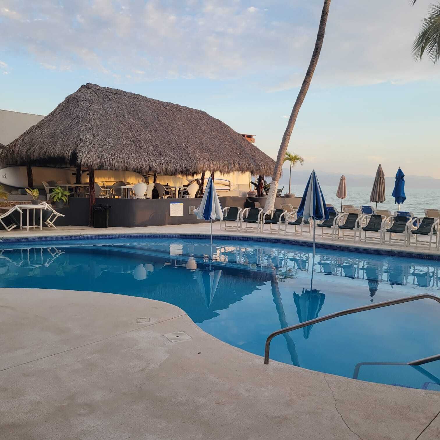 A pool area with lounge chairs, umbrellas, and a thatched-roof structure.