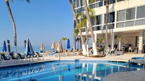 Une piscine d’un bleu éclatant, bordée de chaises longues et d’ombrelles, entourée de palmiers et d’un bâtiment aux grandes fenêtres.