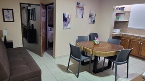 A living room with a brown sofa, a dining table with four chairs, and a kitchen area with wooden cabinets and a whiteboard. - Dos recamaras en la playa conveniente (bucerias)