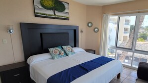 Een hotelkamer met een groot bed, een nachtkastje, een raam met uitzicht op gebouwen en een palmboom, en een schilderij van een boom aan de muur.