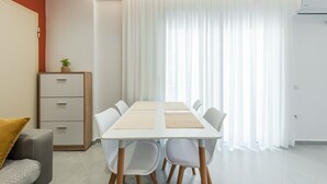 Uma sala moderna com uma mesa de jantar branca, cadeiras brancas, um sofá cinzento e um armário de madeira com uma planta.
