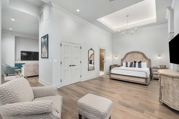 Un dormitorio espacioso con una cama grande, una zona de estar con una butaca y reposapiés, un televisor y un espejo.