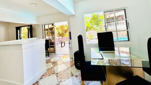 Una habitación amplia con una mesa de comedor de cristal, un sofá y un televisor. Hay un ventanal con vistas a un jardín y un edificio.