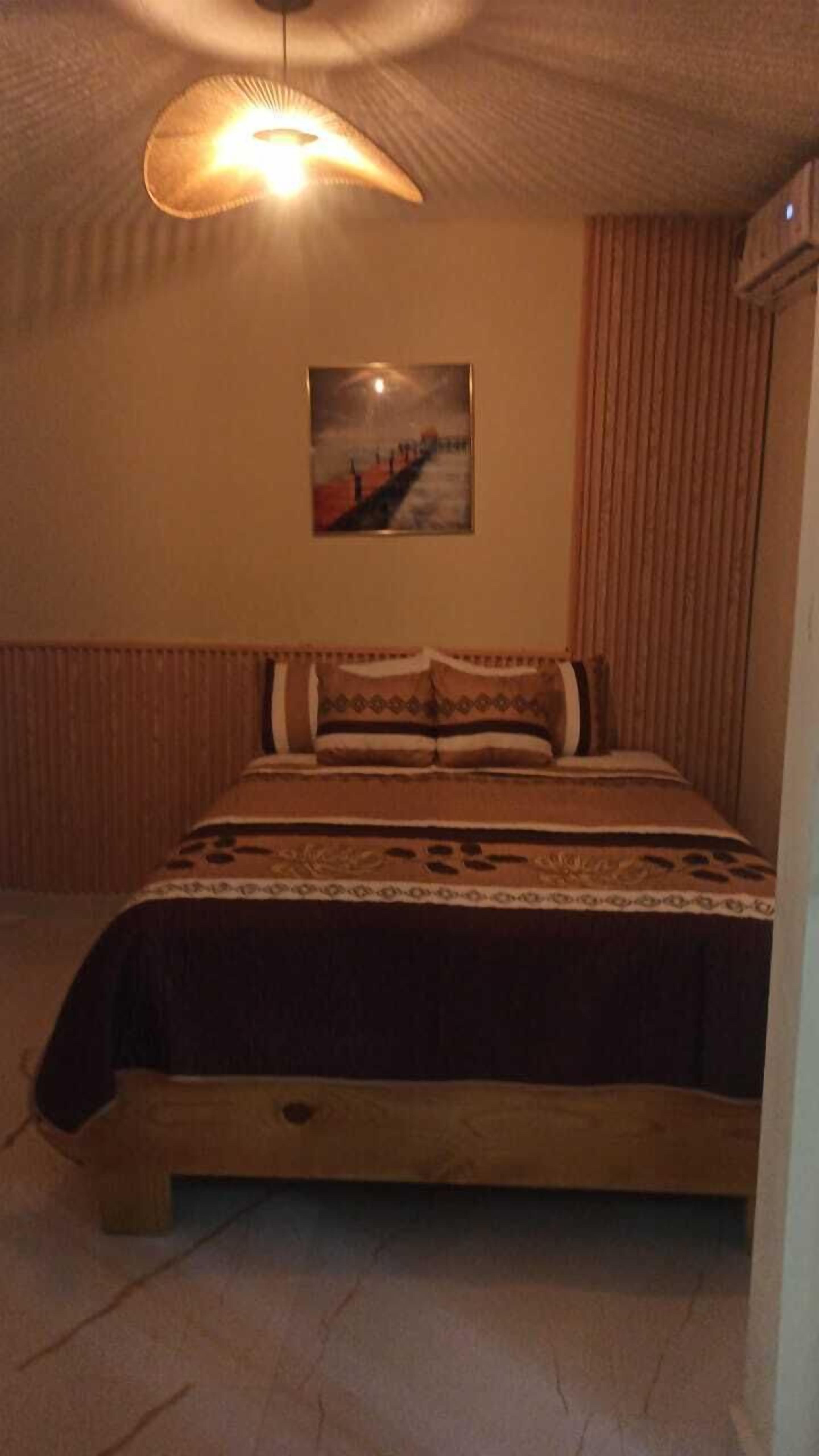 Ett sovrum med en säng, en hängande lampa, en tavla på väggen och ett fönster med gardiner.