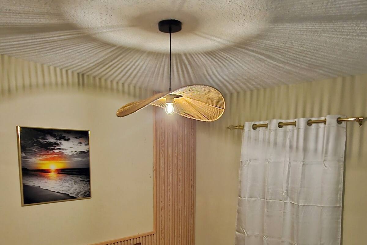 Un dormitorio con cama, ventilador de techo, lámpara de pared, ventana con cortinas e imagen en el marco en la pared.