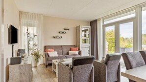 Ein modernes Wohnzimmer mit einer Couch, Sesseln, einem Couchtisch und einem Fernseher. Ein Fenster bietet Blick auf Grünflächen.