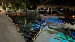 Un complexe hôtelier nocturne doté d’un chemin bien éclairé, de palmiers et d’une structure à toit de chaume.