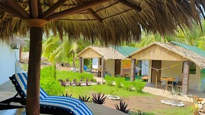 Un complexe hôtelier doté de bungalows aux toits de chaume, d’un espace détente avec des coussins à rayures et d’un jardin bien entretenu.