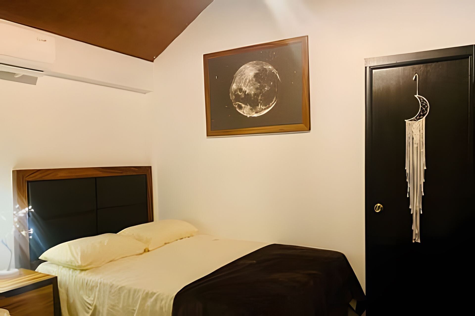 A bedroom with a bed, a wall-mounted air conditioner, a framed picture of a moon, and a door with a towel hanging on it.