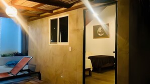 Una stanza con un letto, un tavolo e una sedia. Si vede una finestra e un ingresso che porta in un'altra stanza.