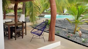 Un'area salotto esterna coperta con tavolo e sedie, un'amaca e vista su una piscina e piante tropicali.