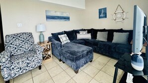 A living room with a dark blue sofa, a patterned armchair, a wooden side table with a lamp, and a ceiling fan. - Two Bedroom One Bath Beachfront Condo (South Padre Island)