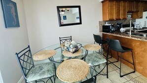 A dining area with a glass table surrounded by chairs, a kitchen area with wooden cabinets, and a mirror on the wall. - Two Bedroom One Bath Beachfront Condo (South Padre Island)