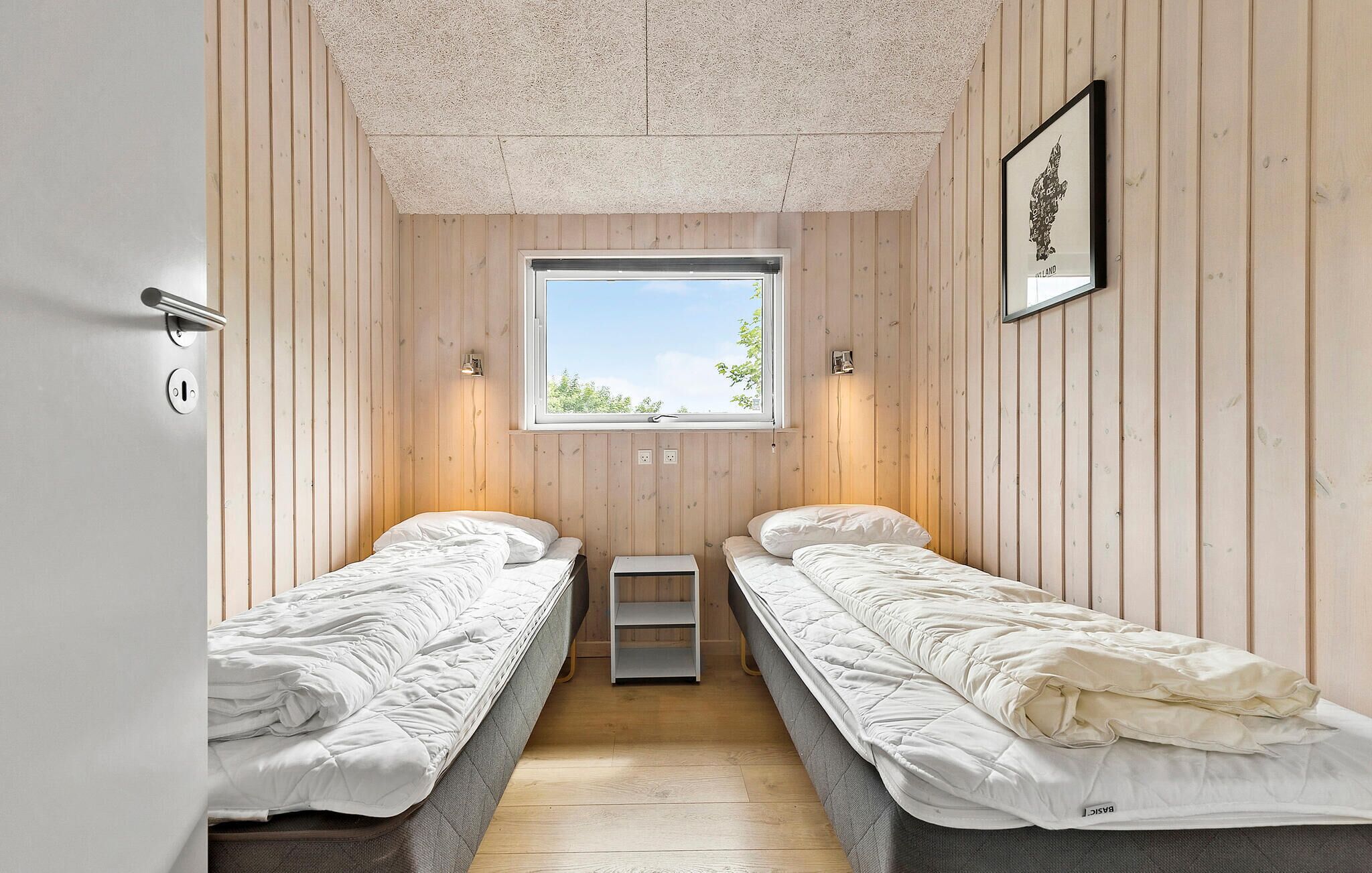 A small, cozy room with two single beds, a window with a view of greenery, and a framed map on the wall.