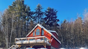 Una cabaña roja con terraza de madera y un ventanal grande, rodeada de nieve en el suelo y árboles.