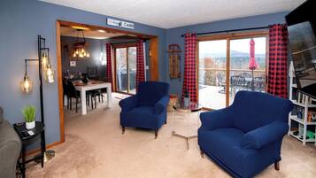 Un salon avec deux fauteuils bleus, un coin repas avec une table et des chaises, et une vue sur une terrasse extérieure, accessible par une porte vitrée.