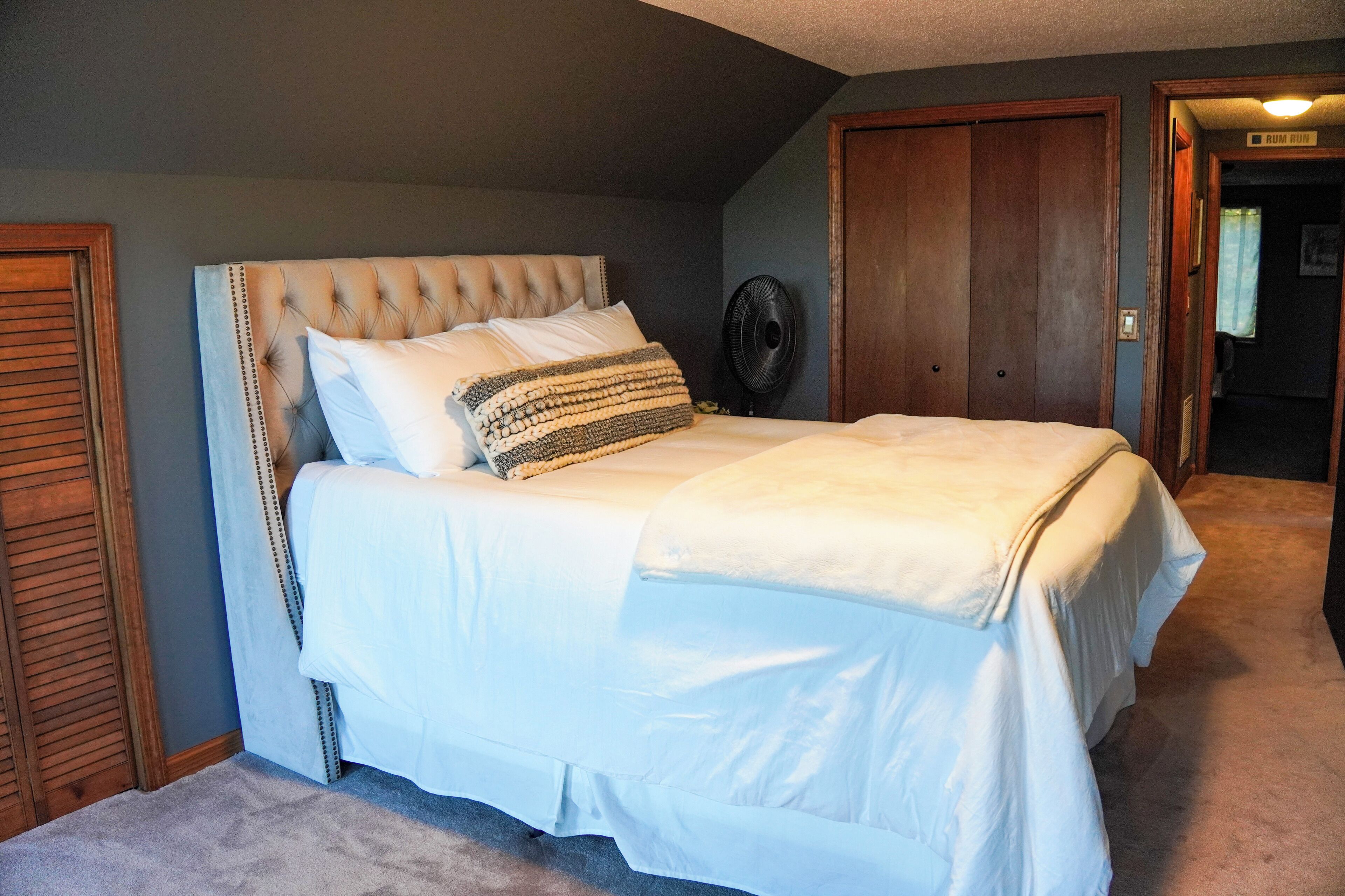 Un dormitorio con cabecera capitonada, cama con ropa de cama blanca, un ventilador y una puerta de madera.
