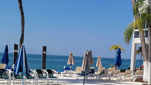Et poolområde med liggestole og parasoller, en klar, blå pool og udsigt til havet.