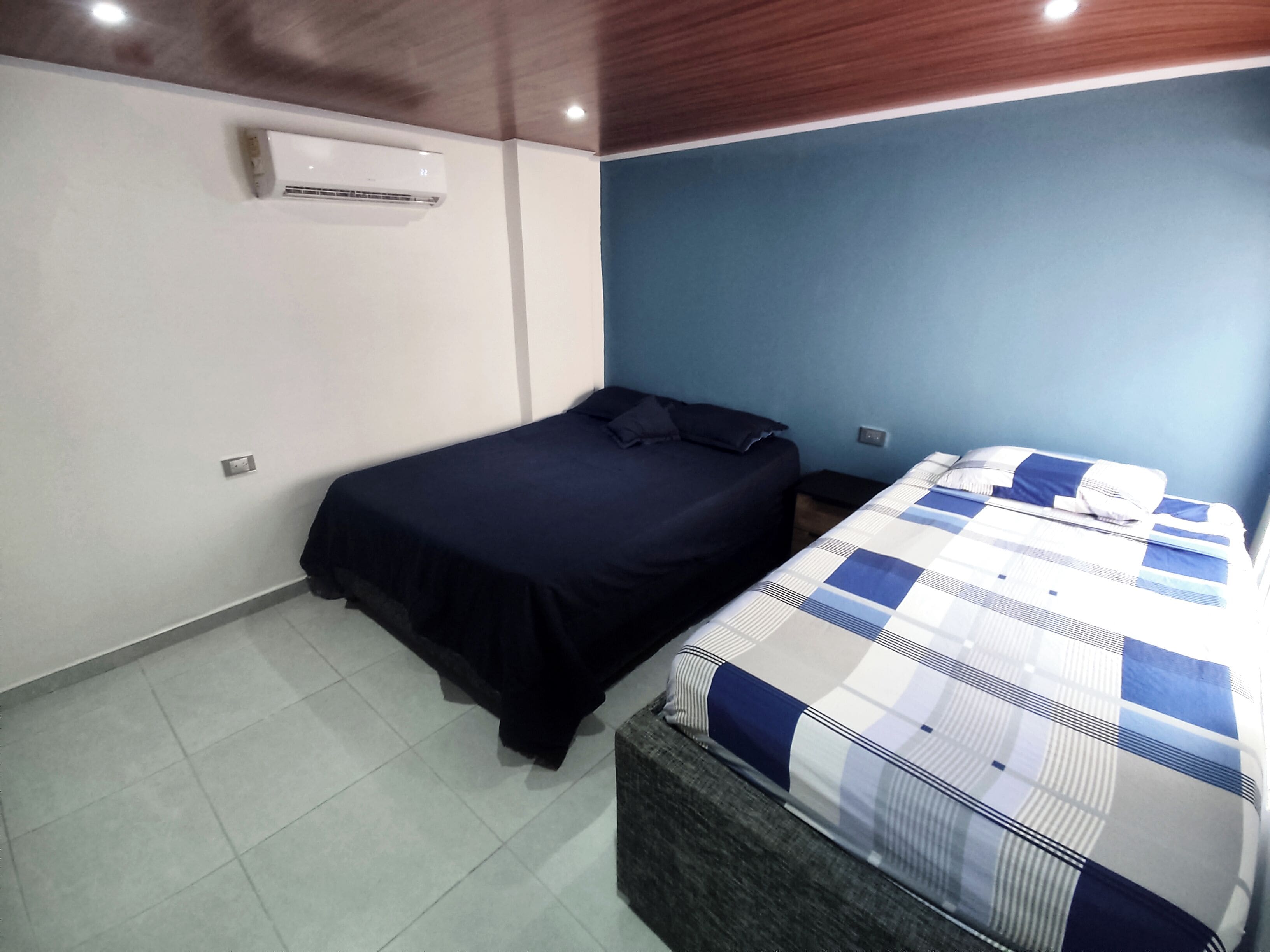 A bedroom with a bed, a bedside table, a nightstand, a wall-mounted air conditioner, and a tiled floor.