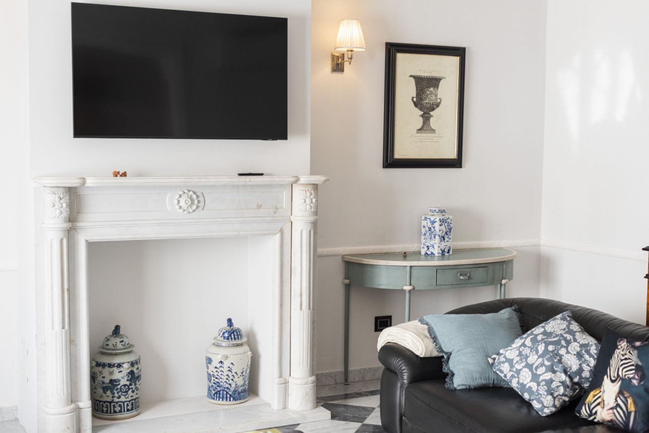 A living room with a fireplace, a mounted TV, a framed artwork, a side table with a vase, and a sofa with patterned cushions.