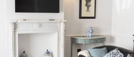 A living room with a fireplace, a mounted TV, a framed artwork, a side table with a vase, and a sofa with patterned cushions.